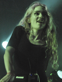 A woman with long wavy bleached hair wears a black shirt and looks excited performing behind a microphone as pale green light illuminates her.