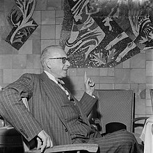 George Szell, seated, in a pin-stripe suit, gesturing with his left hand