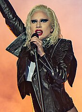 A blonde woman with wet-looking hairstyle singing to a microphone on stage. She is wearing a black leather jacket.