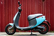An electric motor scooter with a blue body, sitting on its kickstand in front of a red corrugated metal wall.