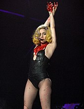 A pale-skinned woman holding her hands crossed and intertwined in the air. She has yellow hair and wears a low-cut bodysuit. Her chest is marked with red liquid.