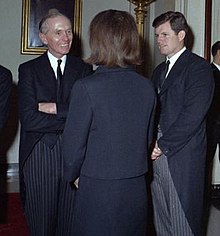Three figures standing and conversing. Jacqueline Kennedy wears a navy dress and has her back to the camera; she is flanked by Douglas-Home (left) and Edward Kennedy (right), both in morning dress.