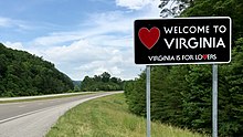 A large rectangular metal sign, mostly black, with the words "Welcome To Virginia" and "Virginia is for lovers" with a red heart symbol on the left stands to the right of a rural road through green hills.
