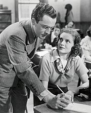 Woman seated at a desk, being instructed by a man, crouching