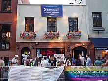A two-story building with brick on the first floor, with two arched doorways, and gray stucco on the second floor, off of which hang numerous rainbow flags.