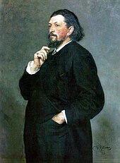 A middle-aged man with medium-length dark hair and a beard, wearing a dark suit, with one hand in his trouser pocket and the other hand on his chin