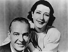 middle-aged white couple smiling at the camera; she is leaning over his shoulder