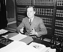 A photograph of Long gesturing with his hands from behind his desk