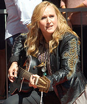 A woman in a black vest and jeans holding a microphone on a stage.