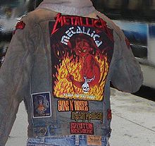 The back of a heavy metal fan wearing a denim jacket is shown. The jacket has patches and artwork for several heavy metal bands attached to the denim. The largest patch is for the band Metallica. It depicts a devil amidst flames.