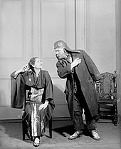 young white woman in Japanese kimono worn as a dressing gown, next to standing middle-aged man in working clothes raising his hand threateningly, ignored by her