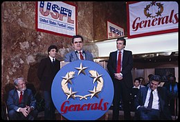 Trump, Doug Flutie, and an unnamed official standing behind a lectern with big, round New Jersey Generals sign, with members of the press seated in the background