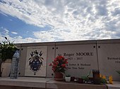 In the middle of a row of white vertical memorials, each about 1 metre wide, and with a horizontal marble shelf before each, a marker with "Sir Roger MOORE", the date "1927–2017", and "Loving Father & Husband", below which is "Our True Saint". Several flower pots and candles are on the shelf for Moore and his neighbours. To the left of Moore's marker is one with a stone plaque labelled "Moore Family", with a coat of arms in blue with gold.