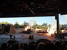 A photograph depicting a replica scene of the Flying Wing being destroyed at a stunt show. Actors portraying Indiana Jones and Marion Ravenwood are positioned in the lower-left corner. The photo is taken from behind audience members.