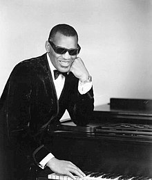 Ray Charles, wearing sunglasses, leaning on a piano from the side with his hand reaching towards the keys.