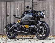 An electric motorcycle with a black body and yellow highlights, sitting on its kickstand in front of a bleached wood wall.