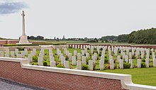 A cemetery of couple hundred white headstones with a terrain of well-manicured grass with plants between each headstone. The cemetery is surrounded by a waist-high red brick wall.