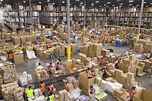 A wide angle shot displays rows upon rows of cardboard boxes full of food. We can see shelves stacked high with even more in the background. Workers are seen in orange vests and masks scattered around the floor packing.