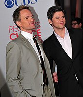A man with dark blonde hair stands next to a man with brown hair as they smile away from the camera