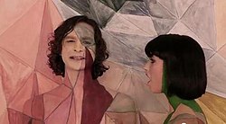 Upper body shot of a man (left) and a woman turned towards him. The man is taller and has dark stragly hair. The woman has dark hair cut in a bob. Both have skin painted in coloured geometric designs. Both are singing. The backdrop has a similarly coloured design closely matching the design on the two people.