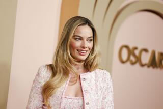 Margot Robbie wearing a pink jacket against a beige and gold backdrop