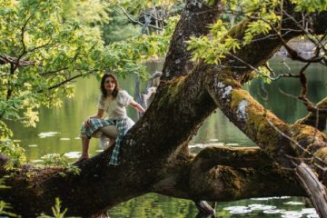 A person sits on a large tree branch which hangs over a lake, with lots of lush greenery in the background