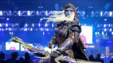 A statue of a character from World of Warcraft with purple skin and pointy ears holding a bow and standing in front of a crowd in a big room with bright lights