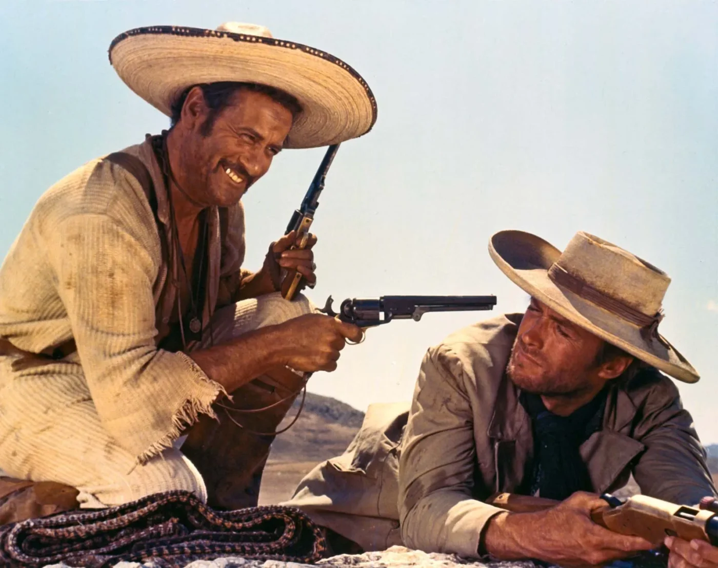 a scene from The Good, the Bad and the Ugly where a man in a hat playfully holds a gun towards Clint Eastwood lying down with a gun poised to the front