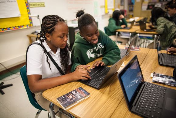 兩位中學女學生在課堂中共用手提電腦。穿著白色上衣的女學生專心看著螢幕，旁邊穿著綠色連帽衛衣的女學生正對著螢幕微笑。