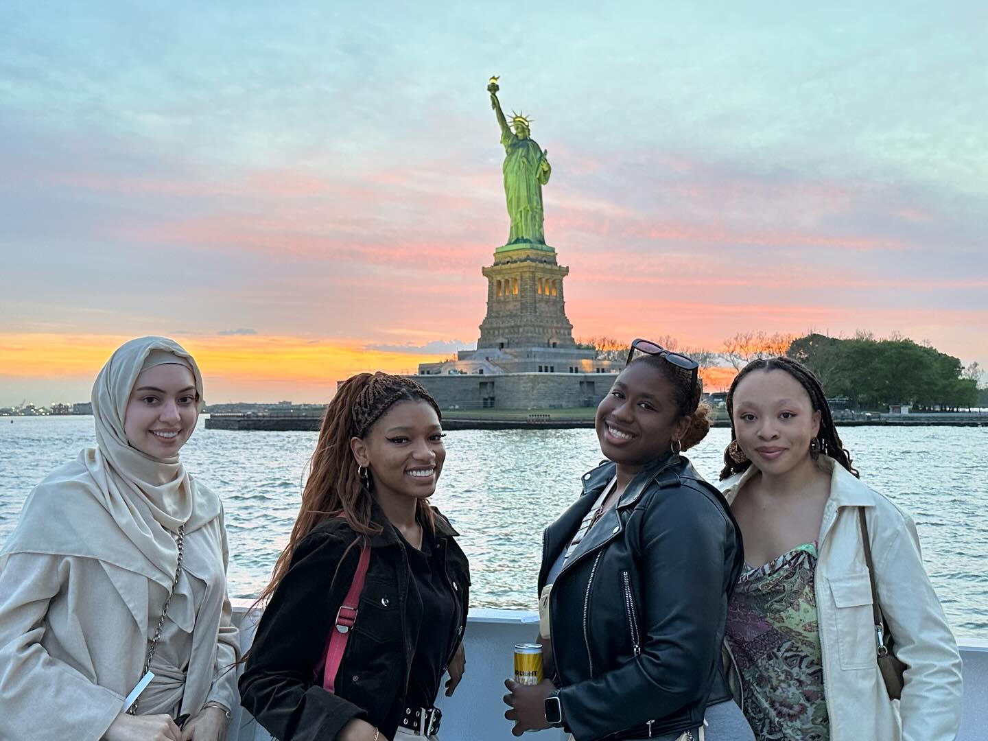Four individuals posing in front of the Statue of Liberty at sunset.