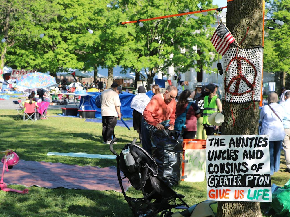 A tree with a sign attached that reads "the aunties and uncles and cousins of greater Princeton give us life." On the same sign is a crocheted blanket with a peace symbol. In the background, people are walking around a green lawn strewn with blankets and tarps. 