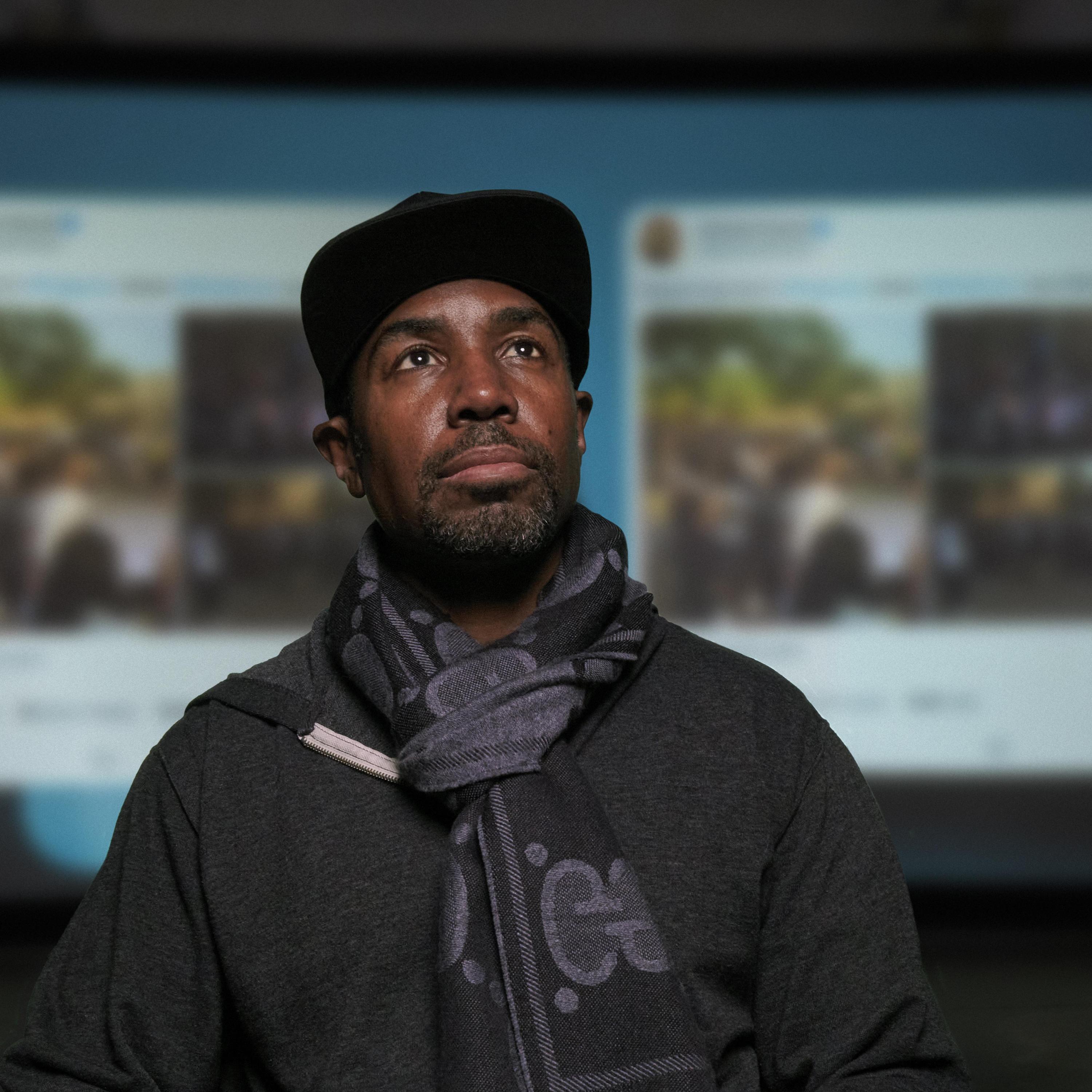 Director Prentice Penny, wearing a scarf and hat and with a screen projecting Tweets in the background.